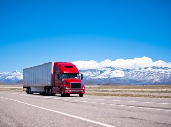 Large beautiful modern classic-modern red truck with a high cab and trailer on a flat stretch of highway on a background of snow-capped mountain ranges, drowning in the clouds and clear blue sky